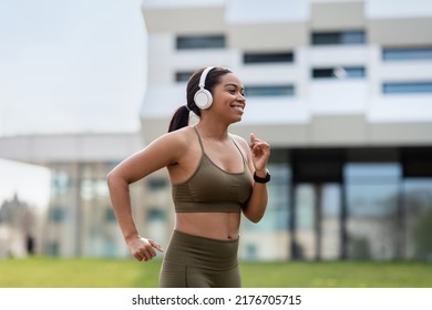 Sporty Young Black Woman Listening To Music In Headphones While Jogging On City Street. Millennial African American Lady Running Outdoors, Enjoying Sports Training In Morning