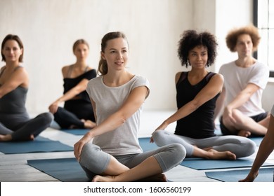 Sporty young diverse students practicing yoga exercise at group lesson, sitting in Parivritta Sukhasana pose on mats, Caucasian and African American people working out in modern fitness center