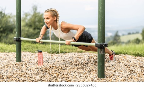 Sporty young female working out calisthenics in an outdoor gym using parallel bars. Beautiful blonde girl with braids works out outdoors doing push-ups on an elevated bar.  - Powered by Shutterstock