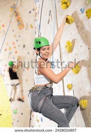 Sporty young girl in safety gear doing difficult wall climb in climbing gym