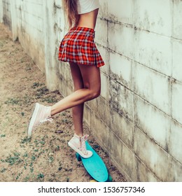 Sporty young lady in a red tartan mini skirt stands on blue penny skateboard trying to look over the fence of a tropical garden. Outdoor lifestyle picture on a sunny summer day.