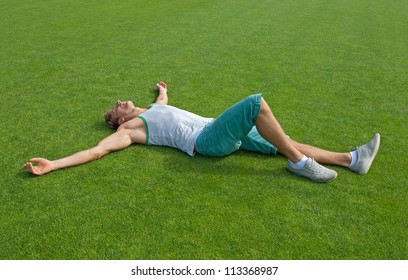 Sporty young man laying on green training field with his arms spread, relaxing.