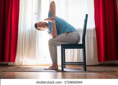 Sporty young man practicing yoga using chair, doing Revolved Chair Pose, Parivrtta Utkatasana, working out at the living room at home
