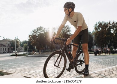 Sporty Young Man Wearing Active Clothes, Helmet And Glasses Sitting On Black Bike Outdoors. Relaxation After Workout. Lifestyles Concept.