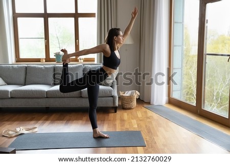 Sporty young slim woman working out in living room, perform Lord of the Dance Natarajasana asana looking out window, boost balance and flexibility, weightloss, yoga training at home, lifestyle concept