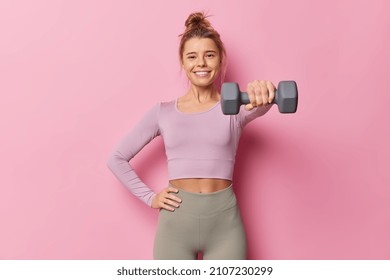 Sporty young woman being in good physical shape keeps hand on waist raises dumbbell has regular fitness training wears sportsclothes poses against pink background. Gym workout and sport concept - Shutterstock ID 2107230299