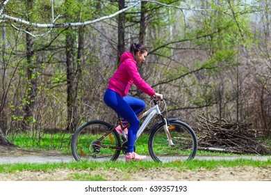 Sporty young woman in a bright pink jacket and jeans rides a bike through the forest on a summer day. Side view