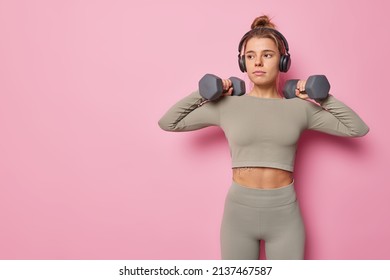 Sporty young woman does exercises with dumbbells bodybuilding workout being in good physical shape dressed in cropped top and leggings listens music via headphones isolated over pink background