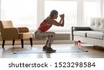 Sporty young woman doing squat morning exercise alone in living room, serious fit girl wearing sportswear crouching training muscles workout at home for healthy body lifestyle concept, side view