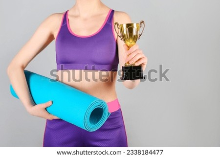 Sporty young woman with fitness mat and gold cup on grey background