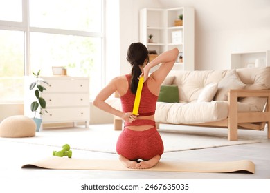 Sporty young woman with stretching band sitting on mat at home, back view - Powered by Shutterstock