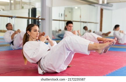 Sporty young woman in white kimono warming up before group martial arts training in gym, doing abdominal crunch.. - Powered by Shutterstock