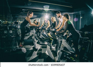 Sporty Young Women And Men Riding Exercise Bikes On Cycling Class With Assistance Of Female Instructor In The Gym.