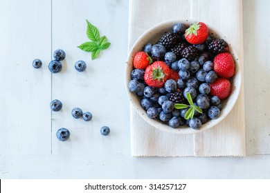 Spotted ceramic bowl with assortment berries blueberries, strawberries and blackberries at white textile napkin over wooden table. Natural day light. Top view - Powered by Shutterstock