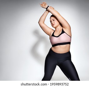 Spoty girl stretches her arms before training. Photo of model with curvy figure in fashionable sportswear on grey background. Sports motivation and healthy lifestyle