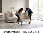 Spouses carrying modern armchair, placing furniture relocating into new home. Happy homeowners 35s couple enjoy moving day, start new life at own house. Fashionable apartment owners, bank loan concept