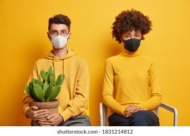 Spouses couple pose at chairs with potted cactus stay at home during self isolation spend time in cozy home during coronavirus quarantine wear face masks feel bored isolated on yellow background - Powered by Shutterstock