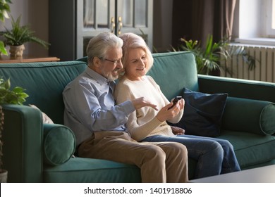 Spouses sitting on sofa grey haired husband help to wife with electronic device cellular usage, elderly couple discuss new application browse internet together older generation and modern tech concept - Powered by Shutterstock