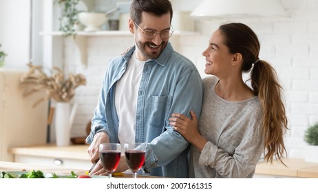 Spouses standing in modern kitchen, cheery wife looks at beloved husband while he cuts vegetables for salad, happy couple enjoy romantic date with red wine. House-warming celebration, new home concept - Powered by Shutterstock