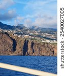 A sprawling coastal cityscape is set against a magnificent backdrop of mountains, with a bright blue sky stretching overhead, blending nature and urban life - Funchal, Madeira, Portugal.