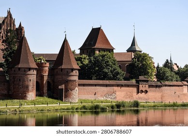 A sprawling medieval castle complex featuring imposing brick towers and walls, set against a clear blue sky with lush green trees and a calm body of water in the foreground