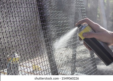 Spray Can With Paint In Hand. Black Paint Is Sprayed From The Spray Can On The Metal Net. Workman Working With Paint Painting