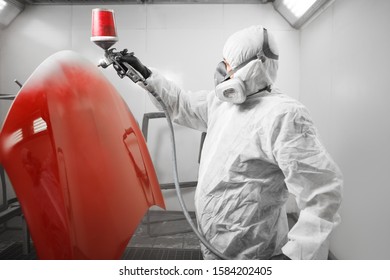 Spray painter worker in protective glove with airbrush pulverizer painting red car body element in white paint chamber.