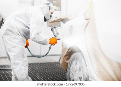 Spray painter worker in protective glove with airbrush pulverizer painting car body in white paint chamber.