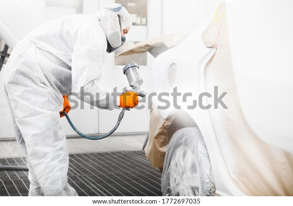 Spray painter\
worker in protective glove with airbrush pulverizer painting car\
body in white paint\
chamber.