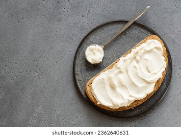Spread cheese (ricotta, mascarpone, philadelphia or cream cheese) swirls on slice of oat wholegrain bread with tea spoon. Top view. Copy space. Grey background. Sandwich or toast on plate.