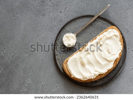 Spread cheese (ricotta, mascarpone, philadelphia or cream cheese) swirls on slice of oat wholegrain bread with tea spoon. Top view. Copy space. Grey background. Sandwich or toast on plate.