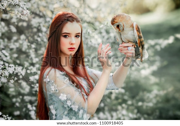 Spring Beautiful romantic red haired girl in blue lace dress standing in blooming garden with owl. Dreaming young model looking at camera. Fantasy art work.