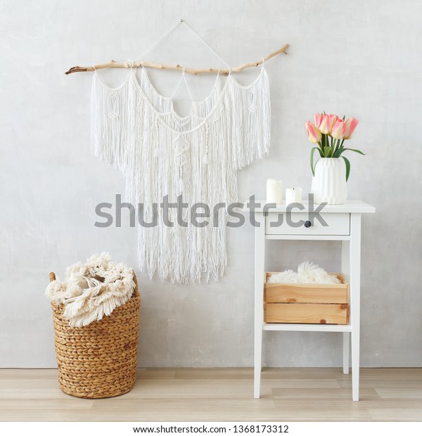 Spring boho home interior decor: macrame wall
hanging decoration, white bedside table, ceramic vase with bouquet
of pink tulips flowers, candles, wicker basket, plaid. Light cozy
modern stylish room.