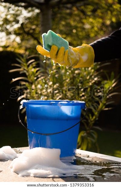 Spring Cleaning outside with big yellow cleaning\
gloves, water, soap and a big blue bucket with soap. Cleaning the\
table. Soap Foam on the table. Hands in bucket. Holding and\
wringing cleaning\
sponge.