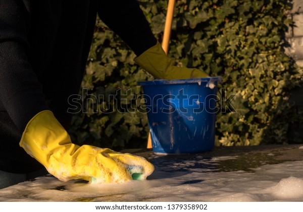 Spring Cleaning outside with big yellow cleaning\
gloves, water, soap and a big blue bucket with soap. Cleaning the\
table. Soap Foam on the table. Hands in bucket. Holding and\
wringing cleaning\
sponge.