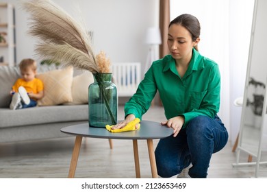 Spring Cleaning. Young Mom Dusting Off Coffee Table And Tidying Up In Living Room While Her Toddler Son Watching Cartoon On Cellphone, Sitting On Sofa On Background