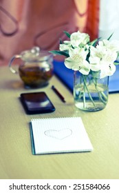 Spring composition for St. Valentines Day: bouquet of white freesia flowers, smartphone, pen and notebook with painted heart. Selective focus. Blurred multicolored background. Vertical vintage image