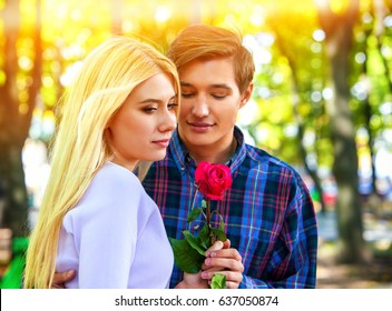 Spring Couple Walking And Kissing Summer Park. Boyfriend And Girlfriend Walk In Sun Shine. First Date Of Two Young People . Guy Gives Girl Red Rose Flower. Fairy-tale Love At First Sight.