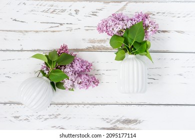 spring decorative composition of two white ceramic mini vases with branches of purple lilac on a white wooden table. Flat original look