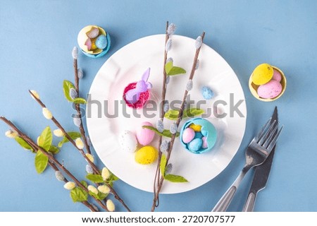 Spring Easter holiday menu table setting, cutlery and plate on pastel blue background with checkered napkin, festive ribbon, Easter festive decor with cute rabbits, Easter chocolate eggs