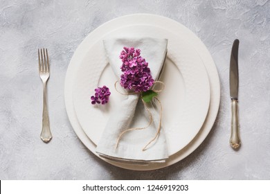 Spring elegant table place setting with violet lilac flowers, silverware on vintage table. Top view.