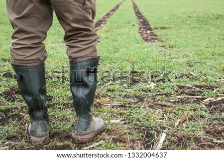 In the spring a farmer stands with his green rubber boots on the edge of his freshly sown field. In the background you can still see the lane of the tractor.