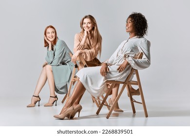 Spring fashion collection. Group portrait of different beautiful young women in elegant dresses sitting on chairs over light background. Concept of beauty, fashion, business, ad.