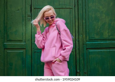 Spring fashion concept: happy smiling fashionable woman wearing trendy pink sport chic style outfit posing in street on green background. Sunglasses, pink hoodie, trousers. Copy, empty space for text - Shutterstock ID 1959024109