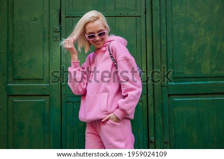 Spring fashion concept: happy smiling fashionable woman wearing trendy pink sport chic style outfit posing in street on green background. Sunglasses, pink hoodie, trousers. Copy, empty space for text
