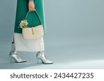 Spring fashion details. Trendy white leather bag with snowdrops in elegant outfit. Woman wearing green coat, skirt, silver ankle boots, posing on blue background. Copy, empty, blank space for text