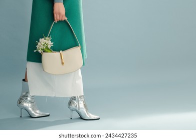 Spring fashion details. Trendy white leather bag with snowdrops in elegant outfit. Woman wearing green coat, skirt, silver ankle boots, posing on blue background. Copy, empty, blank space for text