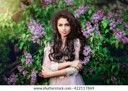 Spring fashion girl outdoors portrait in blooming trees.Beauty romantic woman in flowers of lilac.Sensual lady with long dark shiny hair. Beautiful girl in lacy dress looking at camera. Warm photo.