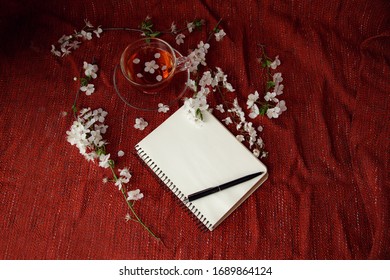 Spring flat lay top view home office workspace - notebook with tea cup and cherry blossom branches on a red desk background