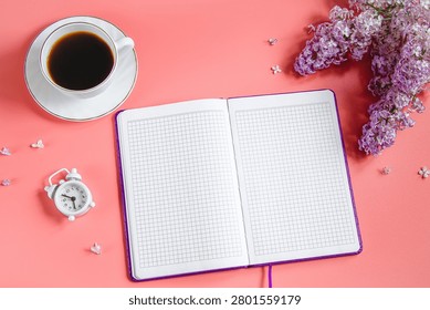 Spring flatlay. Notepad, lilac flowers and cup of coffee on coral background. Early breakfast concept, business plan in office.